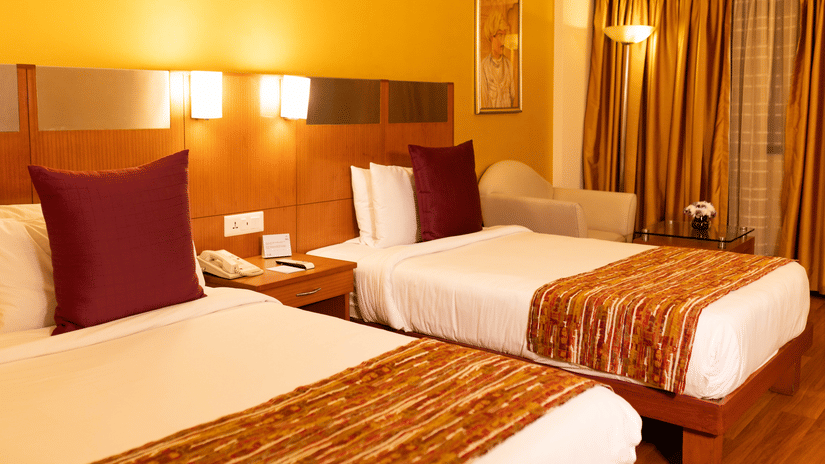 A deluxe room at The Suryaa New Delhi featuring  twin beds with padded headboards, bedside lamps on a shared panel, a window with drawn curtains, and patterned bed runners.