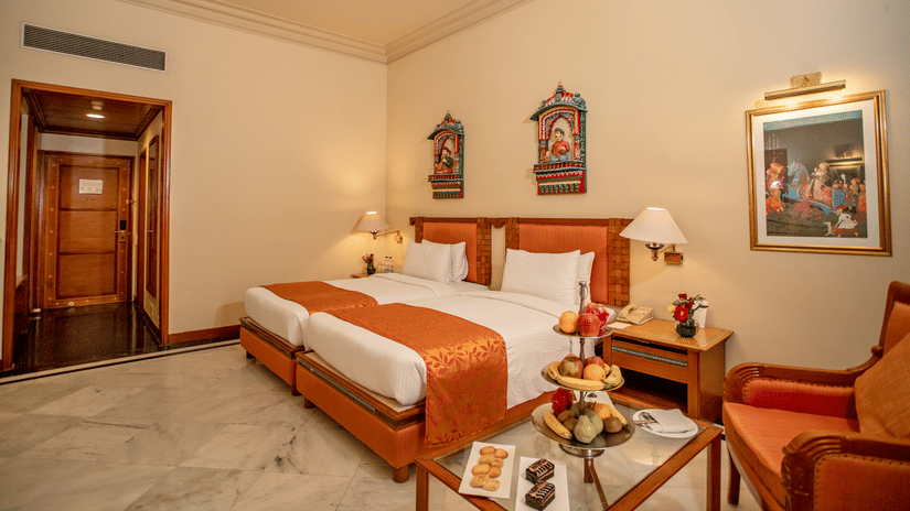 Royal Courtyard Twin Bed with twin beds placed next to each other, a fruit platter on a glass table beside, and traditional art pieces on the walls at The Ummed Jodhpur