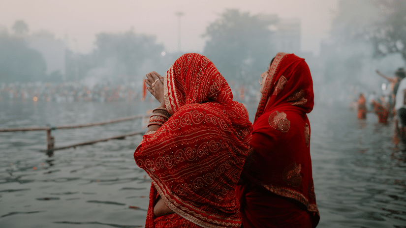 2 women wearing red garments standing in water, with other people visible in the background and a hazy sky above.