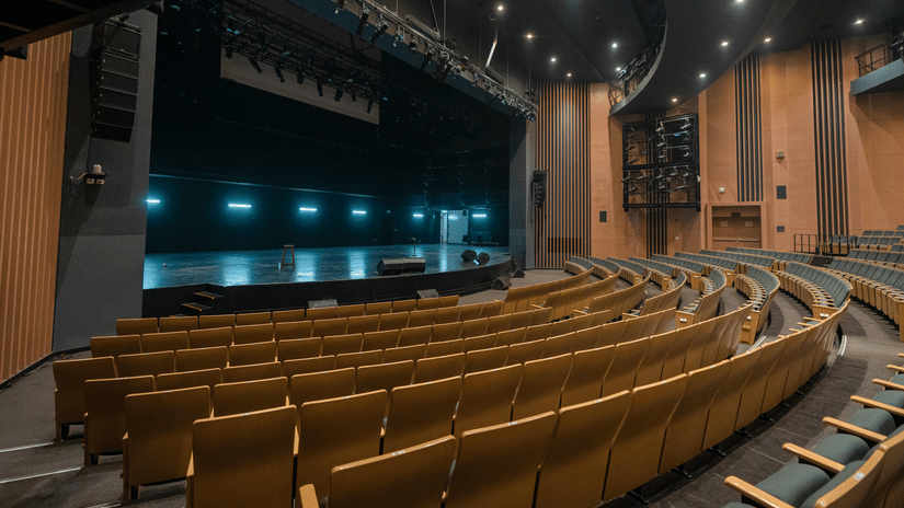 An auditorium with rows of seats facing a stage, overhead lighting, and a large screen at the front.