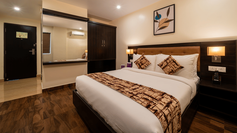 A bedroom interior at VITS Select, Bengaluru, featuring a plush bed, wooden flooring, and a bathroom entrance with mirror.