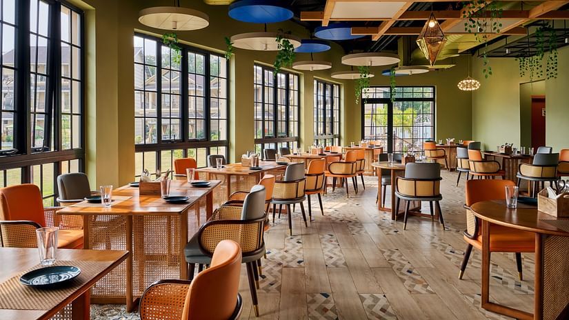 Vibrant and airy restaurant with stylish wooden furnishings, rattan-accented chairs, large windows offering natural light, and a modern ceiling with hanging greenery - Voyage The Solitaire Bliss.