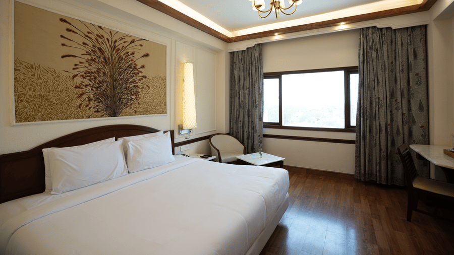 A king size bed with crisp linens sits beneath a floral wall panel and a decorative chandelier in the Deluxe King room at Clarks Amer, Jaipur.