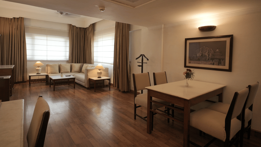 A large living and dining area with wooden flooring, a dining table, upholstered chairs, and wide windows allowing ample natural light in the Executive Suite at Clarks Amer, Jaipur.