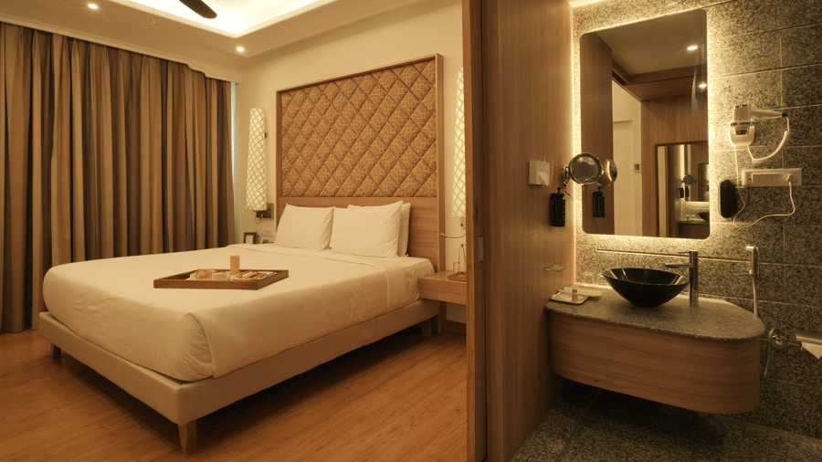 A spacious bedroom with a king size bed, upholstered headboard, wooden flooring, and an attached open bathroom area with a glass partition in the Presidential Suite at Clarks Amer, Jaipur.