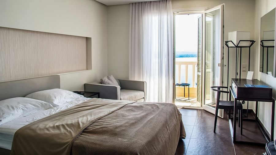 A room with a bed, a small sofa, a desk, and an open balcony overlooking a body of water.