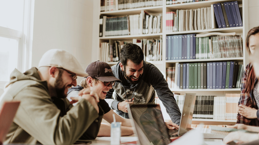 An image of group of people in a library, laughing and working together on a laptop