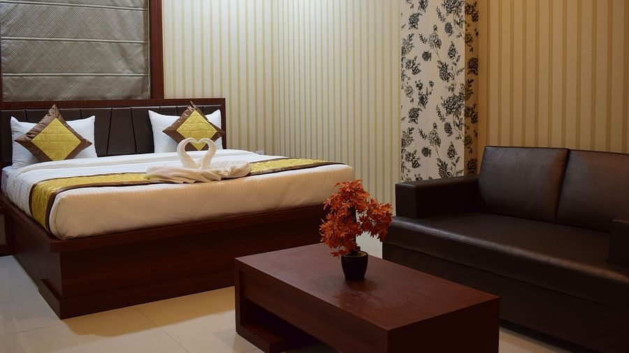A spacious room at Hotel Uday Palace featuring a large bed, a cushioned sofa, and a wooden table with a flower vase.
