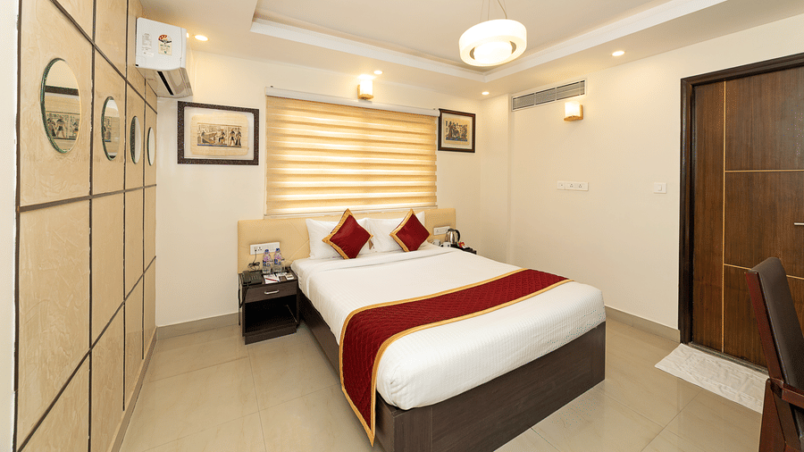 The Deluxe Room of Icon Regency by Bhagini, Whitefield showcasing a double bed with crisp linens, wardrobe, an air conditioning, ceiling lights, closed wooden door, and tiled flooring.