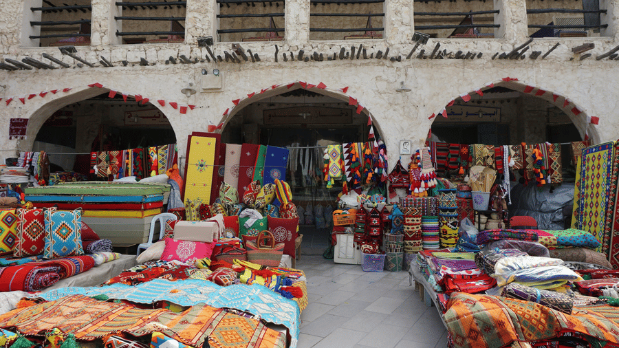 An overview of Souq Waqif market with people selling rugs in a courtyard and an old building in the background.