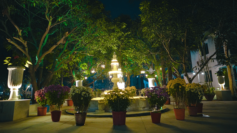 A image of a huge lawn with trees and there is also a fountain surrounded by flower pots - Tangamati Garden Resort