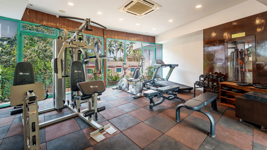 Rosetum Anjuna's air-conditioned fitness center with multi-gym machine, elliptical trainer, treadmill, dumbbell rack, rubber flooring, and garden views through large windows.