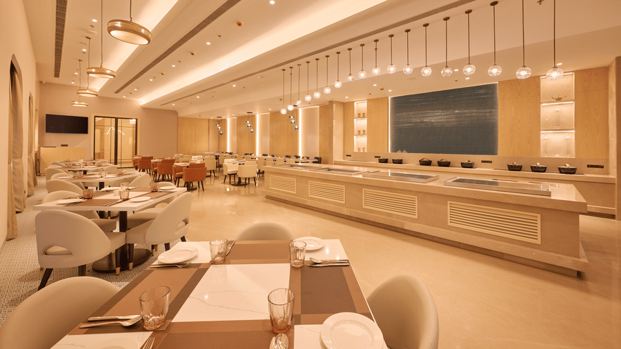 Skywings Sarovar Portico Mathura restaurant dining area with wall-mounted TV, round cushioned chairs, pendant lights, and warm cream-toned interiors.