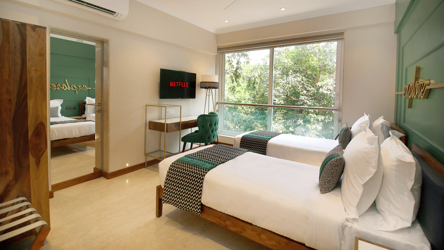 Deluxe Room in Bandra featuring a twin bed with a view outside, accompanied by a wardrobe and mirror on the side, a small table, a chair, and a TV on top at Theory9 - Premium Serviced Apartments