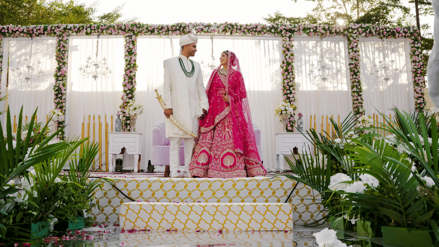 An elegant Indian wedding ceremony with a bride in a pink lehenga and groom in white sherwani standing on a decorated stage.