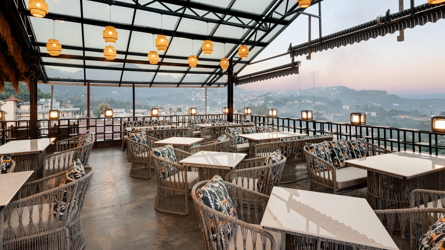 An outdoor rooftop restaurant with a glass and metal roof, woven seating and tables, spherical pendant lights, and a panoramic view of the landscape | Voyage Eastin Suites