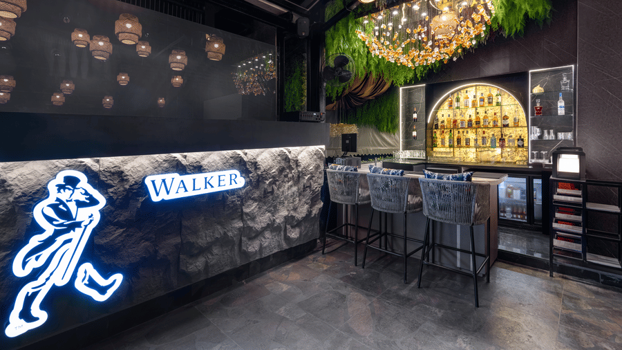 A dark, modern bar area with a lit counter, bar stools, bottles on shelving, and a neon sign of a walking character | Voyage Eastin Suites