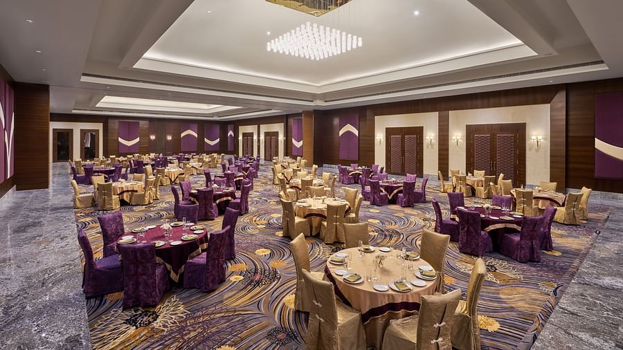 Elegant banquet hall featuring a high ceiling with a modern chandelier, richly patterned carpet, and round tables adorned with gold and purple covers - Voyage The Solitaire Bliss.