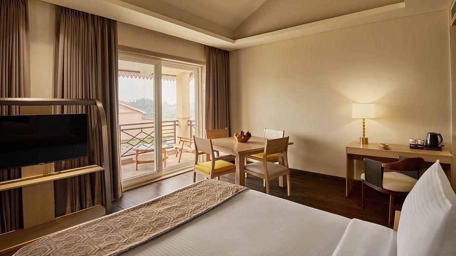 Cosy hotel room with a neatly made bed, flat-screen TV, wooden desk with a lamp and kettle, small dining table with chairs, floor-to-ceiling curtains, and a balcony with a scenic view - Voyage The Solitaire Bliss.