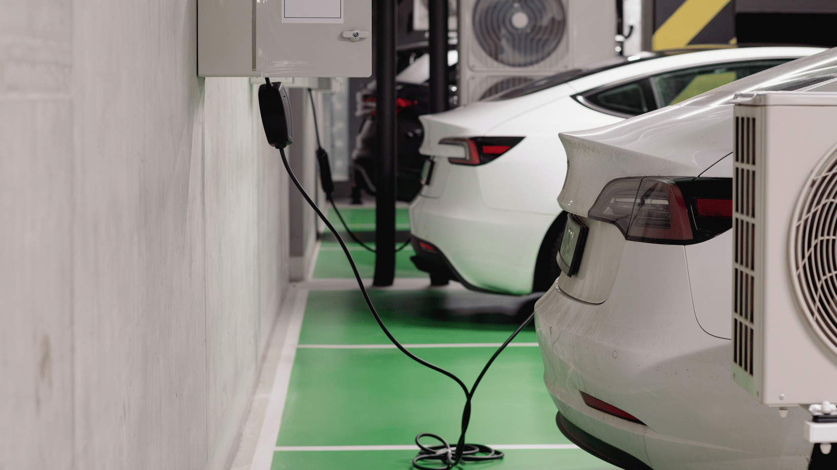 Cars park in a bay. A charger mounts on a wall and connects to a vehicle with a cable. Air conditioners rest on racks near the vehicles.