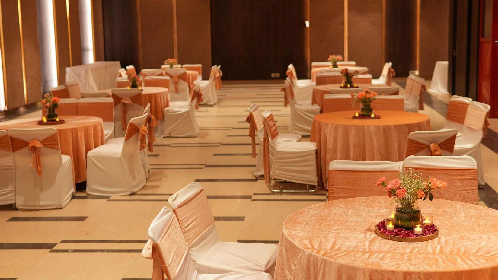 An image of a banquet hall showcasing several elegantly decorated round tables and chairs, illuminated by soft ambient lighting.