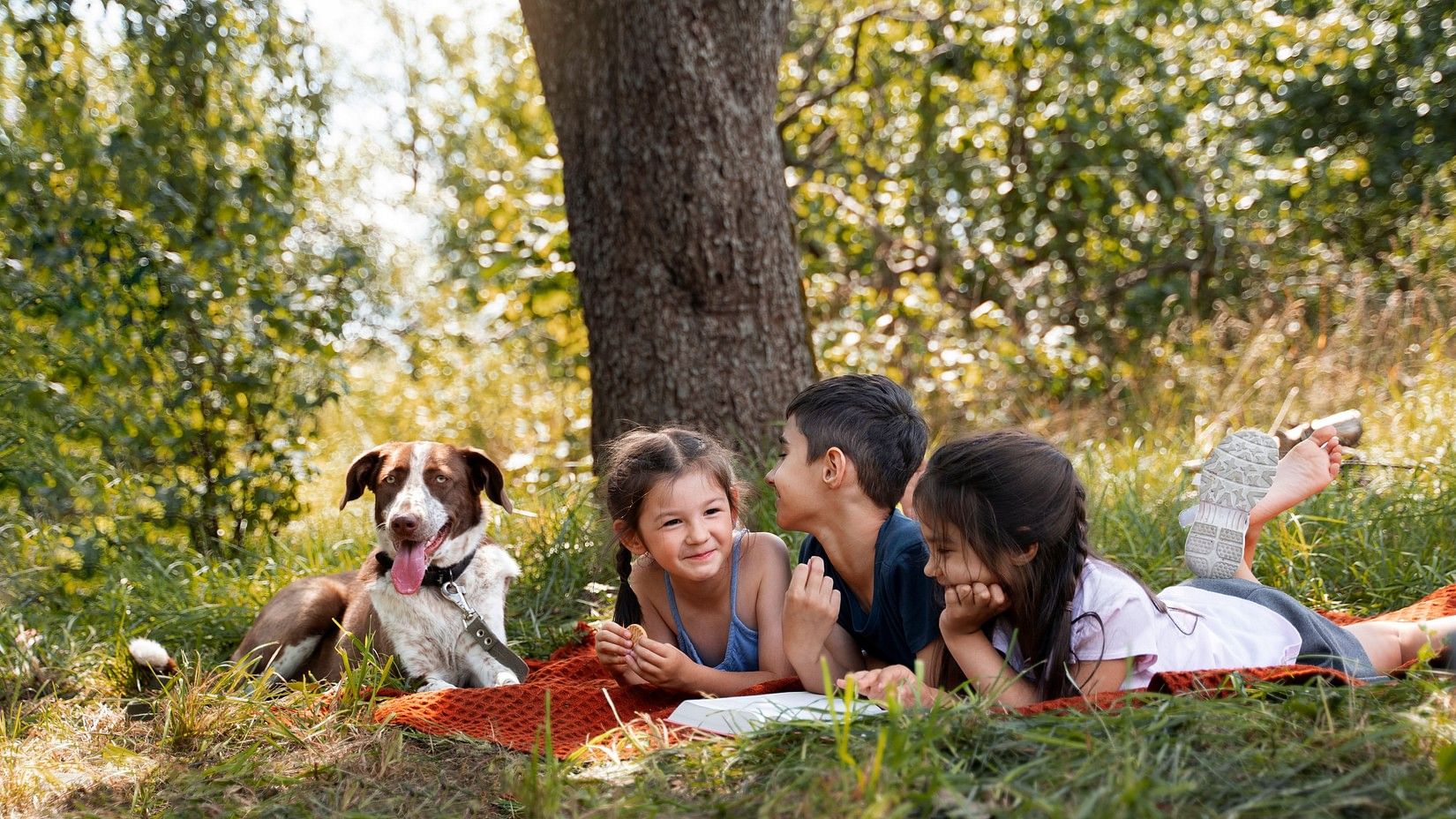 Three children lying on a blanket under a tree, smiling and chatting, with a dog sitting beside them in a grassy outdoor setting.