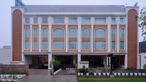 Skywings Sarovar Portico Mathura hotel exterior daytime view with arched windows, cream facade, manicured entrance garden, and overcast sky backdrop.