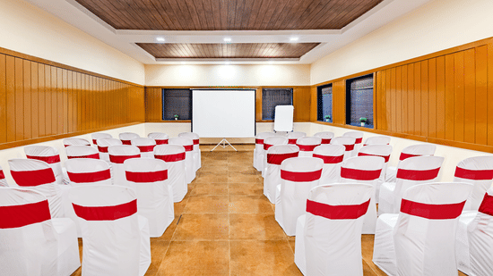 Nature Trails Kudalika conference hall with theatre seating, white and red chairs, projection screen, flipchart, wooden panelling and recessed ceiling lights.