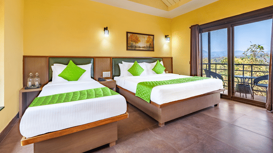 Nature Trails Kudalika river view room with two beds, green bedding, yellow walls, wall lights, framed painting and open balcony with mountain and tree view.