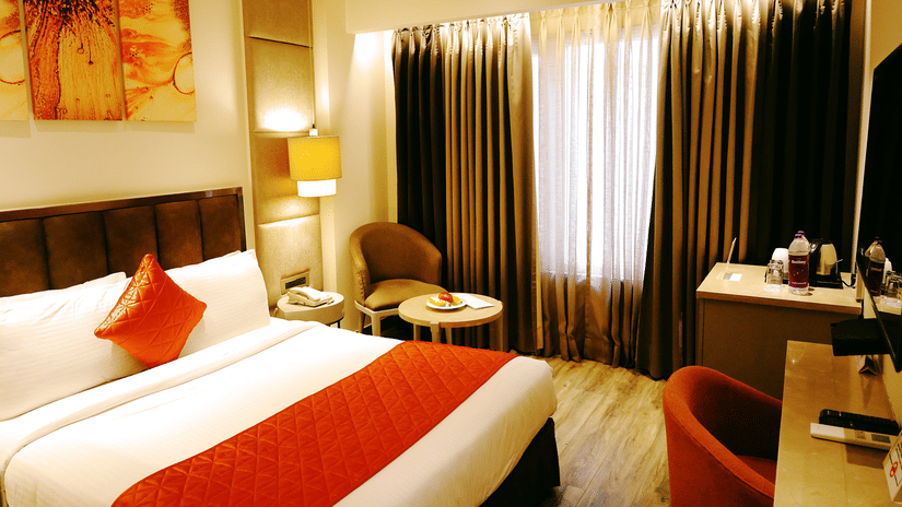 A room at Hotel Saket 27 features a king size bed, upholstered headboard, bedside table, paintings on the wall, desk, chair, television, table lamp and large curtained windows.