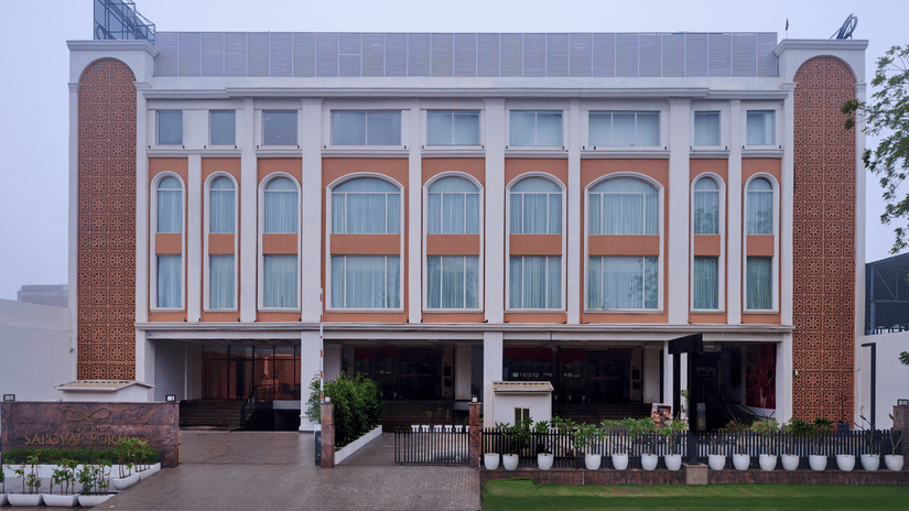 Skywings Sarovar Portico Mathura hotel exterior daytime view with arched windows, cream facade, manicured entrance garden, and overcast sky backdrop.