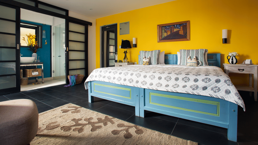 Bright Owl and the Pussycat Hotel in Galle room with a turquoise bed, yellow accent wall, patterned bedding, modern furnishings, and an open bathroom area separated by glass doors.