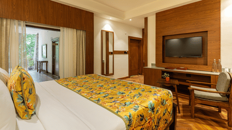A bedroom featuring a bed, a wooden television unit, a chair and a table, a mirror on a wall, and a verandah with trees outside | Summit by the Ganges Beach Resort & Spa, Rishikesh