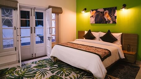 A spacious room at BluSalzz Villas - The Green Roof, Bhimtal, featuring a large bed with a photo frame above the headboard, night lamps glowing on either side, a carpet, and a door leading to the balcony.
