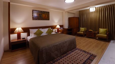 A cosy room with a twin size bed with green and brown accents, bedside tables with lamps, patterned rug, a wall art, a wardrobe, chairs and a wooden table set against green curtains at Denzong Shangrila Lords Inn Gangtok.