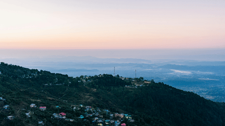 Scenic view of a hillside town with forested slopes, overlooking a vast valley and distant mountains under a pastel sky.