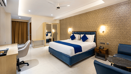 A bedroom at Sunrise A Boutique Hotel, Kondapur featuring a king size bed with cushions, a mirrored wardrobe, a desk and chair, 2 sofa chairs, 2 bedside lamps, and a ceiling fan.
