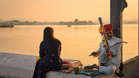 Person sitting by Lake Pichola at sunset with a traditional musician nearby.
