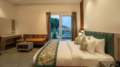 A modern hotel room with a king-size bed, a seating area, and a balcony offering a view of the lush green mountains.