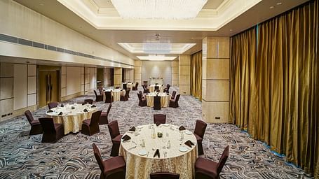 A banquet setup in a large hall with round tables, cushioned chairs, and stylish chandeliers