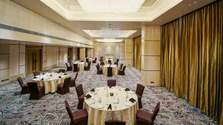 A banquet setup in a large hall with round tables, cushioned chairs, and stylish chandeliers - Grand Madurai by GRT Hotels