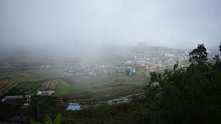 A landscape view of the hills shrouded in mist, with terraced fields and colourful rooftops visible among the greenery.