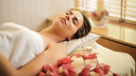 Woman wrapped in a white towel lying on a massage table with her eyes closed. There is a small lit candle and pink flowers nearby.