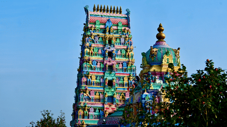 A south Indian temple gopuram featuring multiple tiers with colorful sculpted deities and figures, rising above surrounding trees against a clear blue sky.