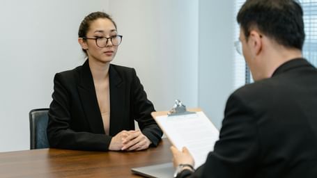 Two professionals with black suits are engaged in a  interview at an office desk with a clipboard. 