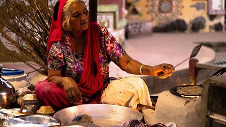 A woman in traditional attire cooks over a fire on a stove, surrounded by cooking utensils and a vibrantly decorated background.