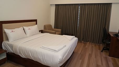 corner view image of the double bed with a huge window in the corner covered with brown curtains - Hotel Grand Sigma