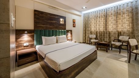 This spacious Maharaja Deluxe Room in Hotel Highway King, Behror, is furnished with a plush king size bed with upholstered headboard, ample lighting in the room, a seating area for two and curtains drawn together.