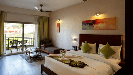 Room featuring green curtains open to a balcony view with 2 chairs kept outside, a large bed, 2 fans, a painting above the bed, and swan shaped towel pair kept at the foot of the bed at Hotel Sonar Bangla, Taki.