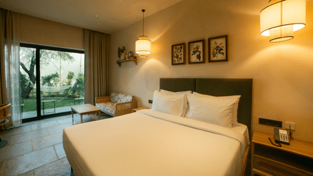 A king size bed sits under 3 framed pictures, with a sliding glass window opening to an outdoor lawn and hanging lamps at Jivana Wellness Retreat, Bhopal.